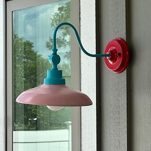May include: A wall-mounted light fixture featuring a bright red circular base, a curved teal arm, and a wide, pale pink dome-shaped shade. The fixture includes a small, central teal connector piece and a single white light bulb visible underneath.