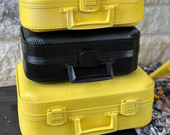 May include: Three stacked vintage-style suitcases. Two are bright yellow, and one is black. Each suitcase has a handle and a clasp closure. The suitcases are made of a textured material, possibly faux leather.