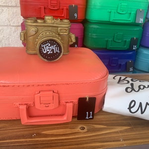 May include: A coral pink vintage suitcase with a gold camera figurine on top. The suitcase has a black tag with the number 11 on it. The camera figurine has a black chalkboard sign with the text "Jez'tu" written on it. The suitcase is sitting on a wooden surface with a white t-shirt with the text "Best day ever" written on it.