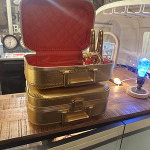 May include: Two gold-colored vintage suitcases stacked on top of each other. The top suitcase is open, revealing a red quilted interior. A gold peace sign statue is inside. The suitcases are on a wooden surface.
