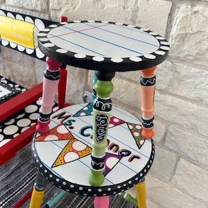 May include: A two-tiered round wooden stool with a white top featuring blue and red lines resembling notebook paper. The legs are painted in various colors including pink, green, orange, and yellow. The lower tier features colorful bunting and the text Ms. Connor and believe.