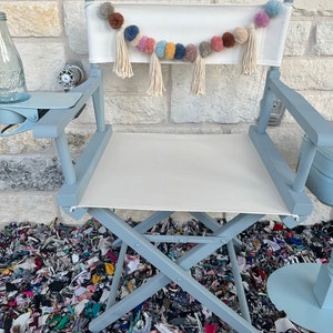 May include: A light blue director's chair with a white canvas seat and back. A colorful pom-pom garland hangs across the back. The chair has a side table attached. The chair is on a surface covered with fabric scraps.