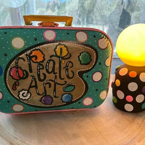 May include: A teal and pink vintage suitcase with a painted palette and the words "Create Art" on the front. The suitcase is sitting on a wooden surface next to a black and white polka dot lamp with a yellow lightbulb.