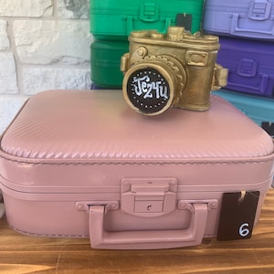 May include: A pink vintage-style suitcase with a gold camera figurine on top. The camera has a black lens with the word "Jez4u" written on it. The suitcase has a brown tag with the number "6" on it.
