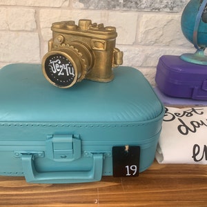 May include: A teal blue suitcase with a gold camera on top. The camera has a black lens with the words "Je t'aime" written on it. The suitcase has a black tag with the number 19 on it.