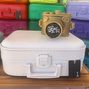 May include: A white suitcase with a gold camera figurine on top. The camera has a black chalkboard with the word "Jez4u" written on it. The suitcase has a black tag with the number "1" on it.
