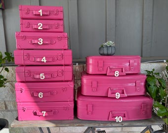 May include: A stack of ten bright pink vintage-style suitcases of varying sizes, each with a white number. The suitcases have handles and clasps. A small succulent in a striped pot sits on top of the stack.