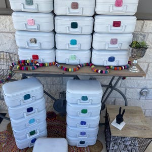 May include: A collection of white vintage-style suitcases with colorful handle accents. The suitcases are stacked and arranged on a wooden table and the floor. A colorful felt ball garland is draped across the table.