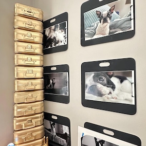 May include: A wall display featuring framed photographs of dogs, alongside a stack of gold-colored miniature suitcases. The photos are in black frames with a unique cutout design. A decorative gold hand statue and a small lamp are also visible.