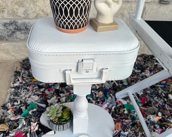 May include: White painted vintage suitcase with a white pedestal base and a white peace sign figurine on top. The suitcase has a decorative gold 'love' inscription on the base. The suitcase is on a colorful rug.