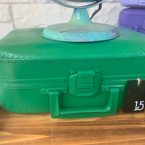 May include: A green vintage-style suitcase with a black tag that says "15" on it. The suitcase has a metal latch and a handle.