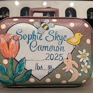 May include: A pink heart-shaped suitcase with a white heart in the center. The heart has the name "Sophie Skye Cameron 2025" written in blue cursive. The suitcase is decorated with a green tulip, a yellow chick, a pink bunny, and white daisies.