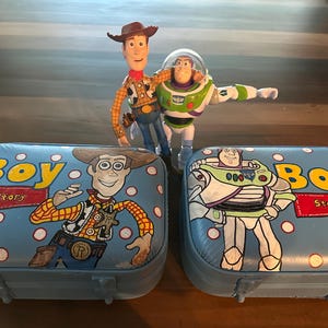 May include: Two blue and white toy storage boxes with a Toy Story theme. One box features Woody, the cowboy doll, and the other box features Buzz Lightyear, the space ranger doll. Both boxes have the words "Boy Story" printed on them.