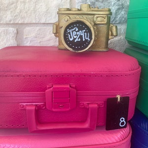 May include: A pink vintage suitcase with a gold camera on top. The suitcase has a black tag with the number 8 on it. The camera has a black lens with the word "Je t'ai" written on it.