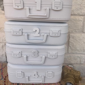 May include: Four white suitcases stacked on top of each other. Each suitcase has a number on the front, from 1 to 4. A gold camera is in the bottom right corner of the image.