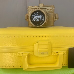 May include: A yellow vintage suitcase with a gold camera on top. The camera has a black chalkboard with the text "Je t'aime" written in white chalk. A black tag with the number "12" is attached to the suitcase.