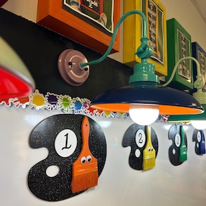 May include: A wall-mounted lamp with a teal arm and a dark blue shade featuring an orange interior. Below, black paint palette cutouts are numbered one through four, each decorated with a colorful paintbrush featuring googly eyes.