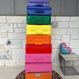 May include: A stack of colorful suitcases and boxes arranged in a rainbow gradient. The top of the stack features a white peace sign sculpture. The bottom of the stack has a black peace sign sculpture. A heart-shaped pillow is on a wooden table.