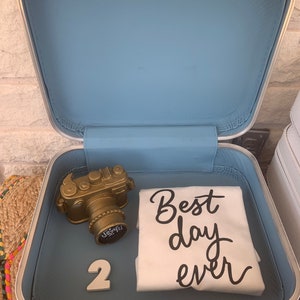 May include: An open white suitcase with a light blue interior. Inside are a gold-colored camera figurine, a white t-shirt with the text "Best day ever" in black, and the number "2".