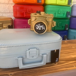 May include: A light blue suitcase with a gold camera figurine on top. The camera has a black chalkboard sign with the text "Jez Tu" written on it. The suitcase has a black tag with the number "2" on it.