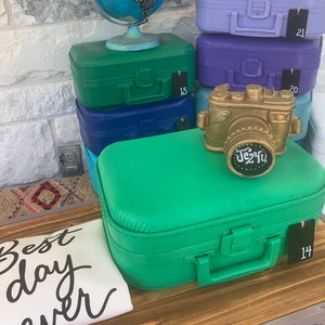 May include: A collection of green, blue, and purple miniature suitcases with a gold camera figurine on top. The suitcases have price tags with numbers on them. The suitcases are on a wooden surface with a white t-shirt that says "Best day ever".