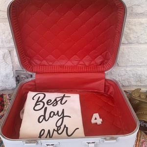 May include: Open white vintage suitcase with a red quilted interior. Inside, a canvas bag displays "Best day ever" in black script, along with a white number "4". The suitcase has silver hardware.