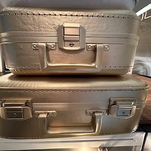 May include: Two gold suitcases stacked on top of each other. The suitcases have a textured surface and are closed. The top suitcase has a lock and a handle.