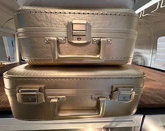 May include: Two gold suitcases stacked on top of each other. The suitcases have a textured surface and are closed. The top suitcase has a lock and a handle.