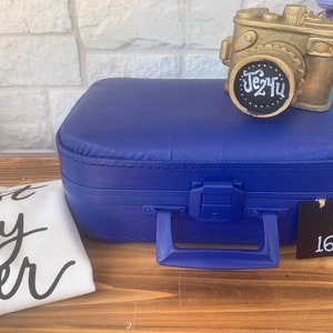 May include: A blue suitcase with a gold camera on top. The suitcase has a tag with the number 16 on it. The camera has a black chalkboard on the front with the text "Je2Hu" written on it.