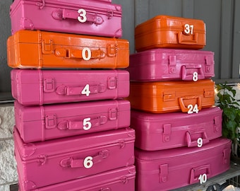 May include: A collection of vintage-style suitcases in shades of pink and orange, stacked on a shelf. Each suitcase has a handle, latches, and a number from 0 to 37. The suitcases are arranged in a decorative display.