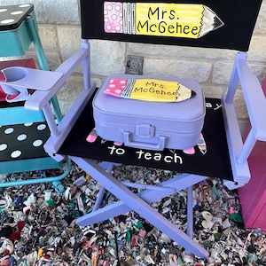 May include: A lavender director's chair with a black seat and backrest. The backrest features a pencil design and the name "Mrs. McGehee". A matching lavender suitcase with a pencil design sits on the seat. The words "to teach" are printed on the seat.