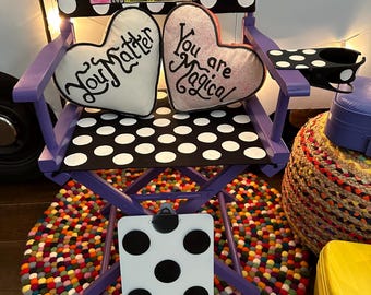May include: Purple director's chair with a black and white polka dot pattern. The chair has two heart-shaped pillows with the text "You Matter" and "You are Magical". The chair is sitting on a colorful round rug with a black and white polka dot clipboard leaning against it.