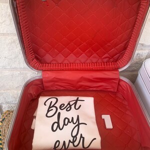 May include: A white vintage suitcase with a red quilted lining. The suitcase is open and a white t-shirt with the text "Best day ever" is inside. The number "1" is also inside the suitcase.