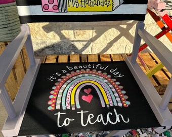 May include: A purple director's chair with a black canvas seat and back. The back is decorated with black and white stripes and a yellow pencil with the text "Mrs. Hernandez". The seat has a rainbow with hearts and the text "it's a beautiful day to teach".