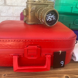 May include: A red vintage suitcase with a gold camera figurine on top. The suitcase has a black tag with the number 9 on it. The camera figurine has the text "Jez4u" on it.