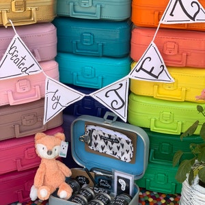 May include: A stack of colorful vintage-style suitcases in gold, pink, blue, orange, yellow, and green. A white bunting banner with the black text Created by J hangs across the front. An open light blue suitcase contains a plush fox, jars, and baby items.