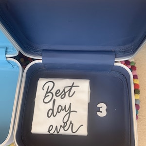 May include: A white t-shirt with the text "Best day ever" in black lettering inside a silver suitcase with a blue interior. The number "3" is written in black on the suitcase.