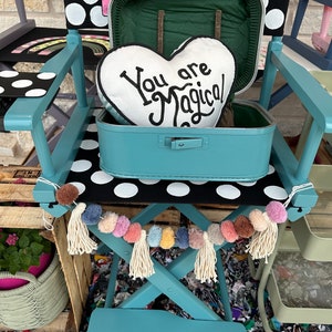 May include: A teal director's chair with a black and white polka dot seat. The chair has a teal suitcase with a heart-shaped pillow that says "You are Magical" inside. The chair is decorated with a white tassel garland with colorful pom-poms.