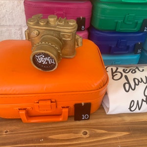 May include: A gold-colored camera figurine sits on top of an orange suitcase with a black handle. The suitcase has a tag with the number 10 on it. A white t-shirt with the text "Best day ever" is lying on a wooden surface.