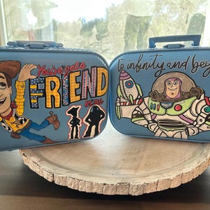 May include: Two blue suitcases with painted designs. One suitcase features Woody from Toy Story with the text "You've got a friend in me". The other suitcase features Buzz Lightyear from Toy Story with the text "To infinity and beyond".