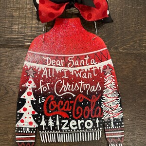 May include: A Christmas-themed wooden decoration shaped like a sweater, featuring the text "Dear Santa, All I want for Christmas is Coca-Cola Zero." The sweater is red, black, and white, with a polka dot bow at the top.