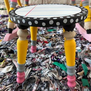 May include: A small round stool with a seat painted to resemble lined notebook paper with a red margin line. The edge of the seat is black with white polka dots. The three legs are painted to look like yellow pencils with grey ferrules and pink erasers.