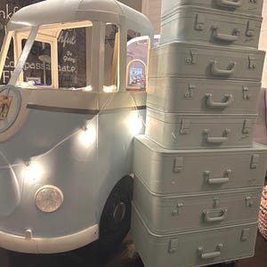 May include: A stack of six vintage suitcases painted in a light blue color. The suitcases have handles and are stacked on top of each other. The suitcases are on wheels and are in front of a blue and white painted Volkswagen bus.