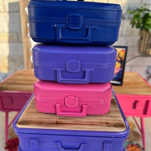 May include: A stack of three vintage suitcases painted in vibrant colors: blue, purple, and pink. Each suitcase has a handle and a clasp. A pair of black button glasses sits atop the blue suitcase. The suitcases are on a purple table.
