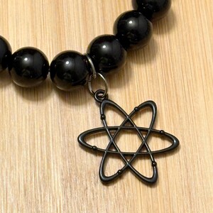 May include: Black beaded bracelet with a dark metal charm in the shape of an atom. The bracelet features large, glossy black beads and a decorative metal clasp. The charm is suspended from a small metal ring.