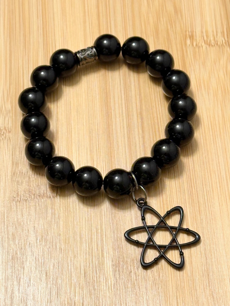 May include: Black beaded bracelet with a dark metal charm in the shape of an atom. The bracelet features large, glossy black beads and a decorative metal clasp. The charm is suspended from a small metal ring.