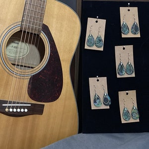 Op de afbeelding: Een display van zes paar druppelvormige oorbellen met een groen, blauw en zwart marmerpatroon. De oorbellen worden gepresenteerd op een zwarte achtergrond met bruin karton achter elk paar.