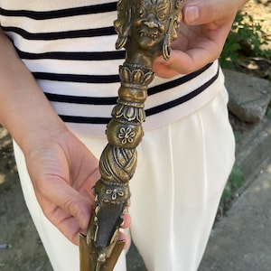 May include: A bronze-coloured ritual phurba dagger featuring a detailed three-faced deity head at the top, a central lotus petal design, a twisted middle section, and a sharp, three-sided blade tip with intricate engravings throughout the metal surface.