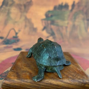 May include: A bronze-colored turtle figurine with detailed shell patterns, positioned on a small, rectangular wooden base. The turtle's legs and head are visible, showcasing its textured surface. The background features a blurred, colorful landscape.