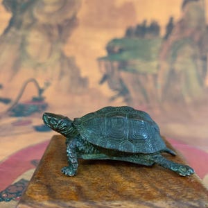 May include: A detailed bronze turtle figurine with a textured shell and detailed legs, resting on a wooden surface. The turtle is a dark green color, and the wooden surface is a light brown. The background features a landscape painting.